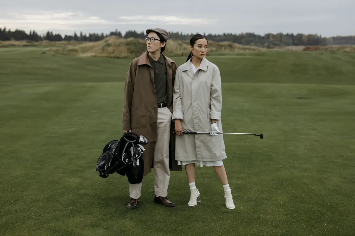Fashionable couple posing on golf course in outerwear, showcasing modern fall fashion trends.